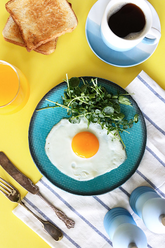 Stock photo showing elevated view of sunny side up fried egg on a turquoise plate served with watercress and cup and saucer of black coffee, glass of orange juice and sliced white toasted bread as a healthy breakfast.
