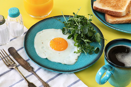 Stock photo showing close-up view of sunny side up fried egg on a turquoise plate served with watercress and mug of black coffee, glass of orange juice and sliced white toasted bread as a healthy breakfast.