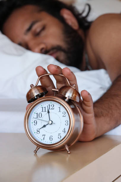 istock Image of Indian man lying on his front in hotel double bed under bedding, hand reaching for night stand to turn off bronze metal case retro alarm clock, double bell, analogue display clock with white dial, focus on foreground 1468720810