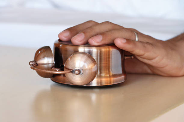 Stock photo showing close-up view of a bronze metal case retro alarm clock with double bell lying face down night stand with hand reaching from under duvet on hotel double bed to turn it off.