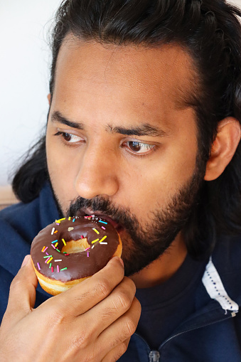 Stock photo showing close-up view of chocolate glazed ring doughnut with sprinkles held by an Indian man ready to take a bite.