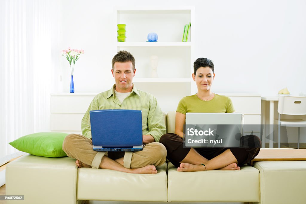 Couple sitting on the sofa and using laptops Young couple sitting on a sofa cross-legged in the living room, using laptops.  20-24 Years Stock Photo