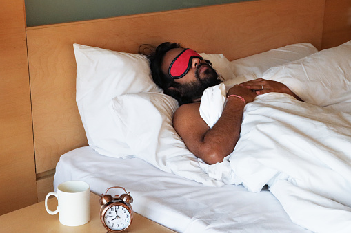 Stock photo showing close-up view of a bronze metal case retro alarm clock with double bell on night stand besides a white coffee mug. An Indian man can be seen lying in the double bed, next to the beside table, with a red sleep eye mask covering his eyes to block out the early morning sunlight.