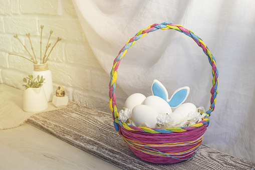 A colorful wicker Easter basket with eggs is on the table in the kitchen. The ears of a hare stick out of the basket. High quality photo