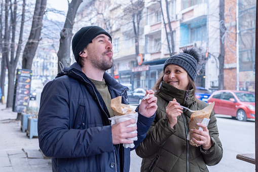 Two cheerful friends, a man, and a woman are eating fast food on the street. They are dressed in warm jackets and hats. They are eating an Arabic doner kebap on the sidewalk in front of the diner and having fun.