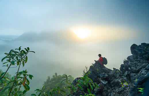 an asian man holding a mobile phone in his hands on the mountain while enjoying a misty sunrise. Adventure tourist landscape photography