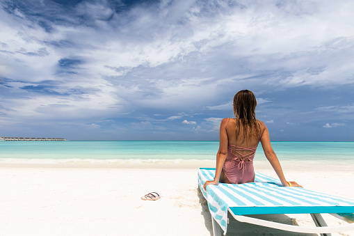 Back view of a woman sitting on a deck chair on the beach and looking at view. Copy space.