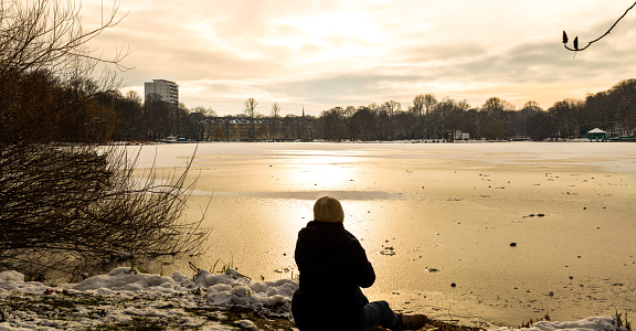 Woman searches for a bit of sunlight in a cold winter day and sits at a frozen lake shore