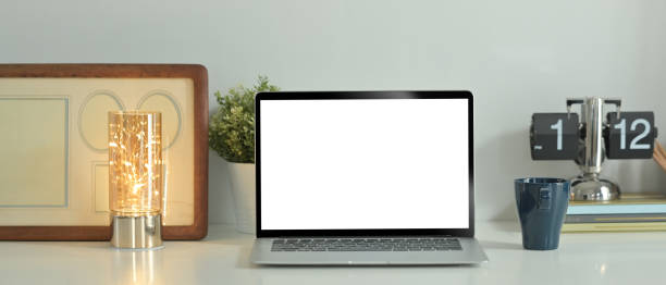 Laptop computer, coffee cup, picture frame and potted plant on white table. Blank screen for text information or content.