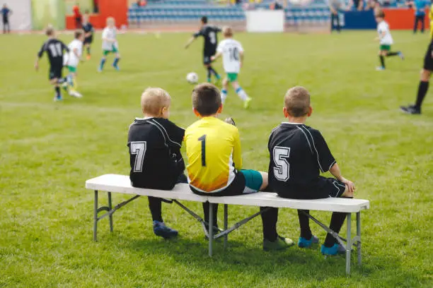 Photo of Young boys in soccer jerseys sitting on the substitute players' bench. Children play sports during school tournaments. Kids having fun attending sports competition