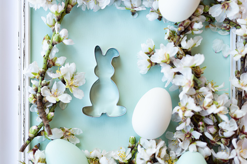 Beautiful flat lay of an easter bunny cookie cutter with almond blossoms on a pastel mint green colored surface with an easter eggs in a vintage picture frame. Without color editing, directly converted from RAW in order to put your own desired preset. Very selective and soft focus. Part of a series.