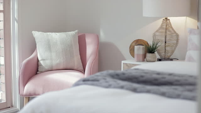 A comfortable pink chair in a pretty bedroom with morning sunlight streaming through the window onto the soft, cozy bed with quilted blankets.