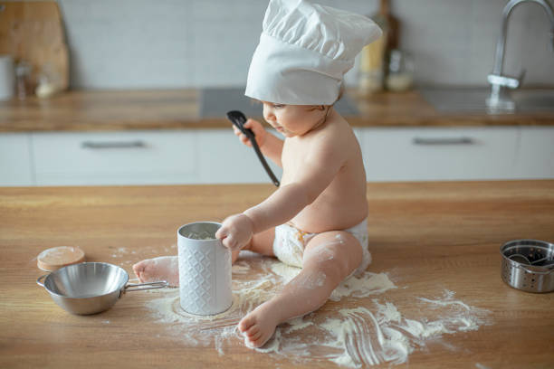 Baby boy in diapers sitting on a kitchen counter wearing a chef's hat , playing with flour and kitchen utensils