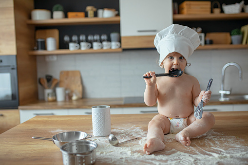 Baby boy in diapers wearing a chef's hat, sitting on kitchen counter, nibbling on kitchen utensils