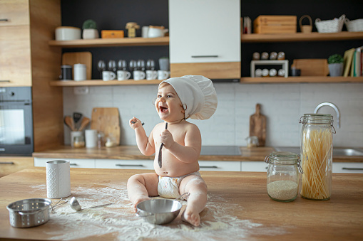 Baby boy in diapers sitting on a kitchen counter wearing a chef's hat , playing with flour and kitchen utensils