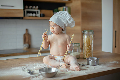 Baby boy in diapers wearing a chef's hat, sitting on kitchen counter, nibbling on spaghetti