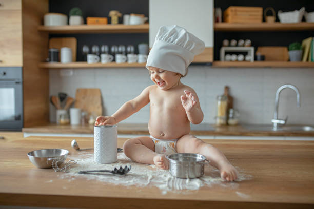 Baby boy in diapers sitting on a kitchen counter wearing a chef's hat , playing with flour and kitchen utensils
