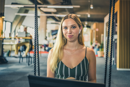 Portrait of smiling young businesswoman using laptop while sitting on swing in office.
