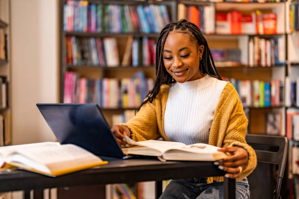 A high school student in a library, studying with a stack of textbooks and a laptop.