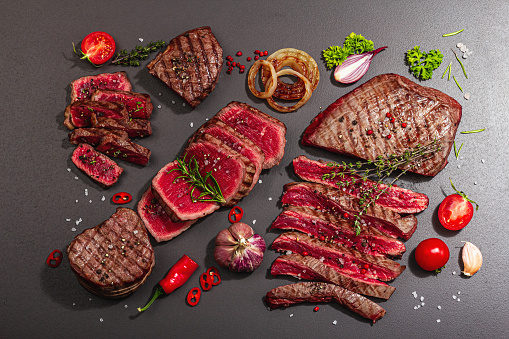 Grilled set of various steaks with vegetables, spices and herbs. Classic meat cuts includes ribeye, eye round, flank and striploin steaks. Black stone background, flat lay, top view