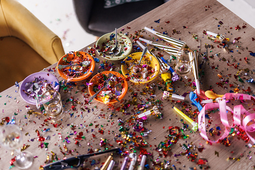 Directly above shot of aftermath of a great celebratory party. Colorful confetti, half eaten cake slices empty glasses, cake fireworks, party horns, balloons and other decor.