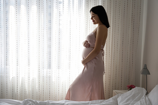 Brunette young woman on second trimester of pregnancy standing by the window in a bedroom. Pregnant female with arms around her belly. Expecting a child concept. Background, cop y space.