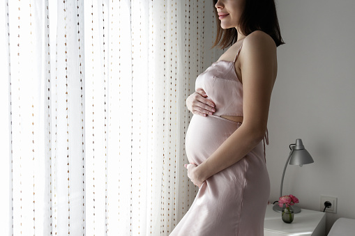 Brunette young woman on second trimester of pregnancy standing by the window in a bedroom. Pregnant female with arms around her belly. Expecting a child concept. Background, cop y space.