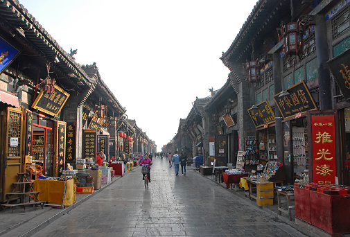 A main street in Pingyao, Shanxi Province, China lined with small shops and stores. A woman is cycling and people are walking.
