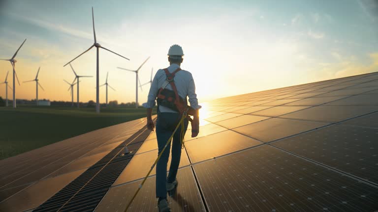 Professional Male Green Energy Engineer Walking On Industrial Solar Panel, Wearing Safety Belt And Hard Hat. Man Inspecting Sustainable Energy Farm With Wind Turbines On Background.