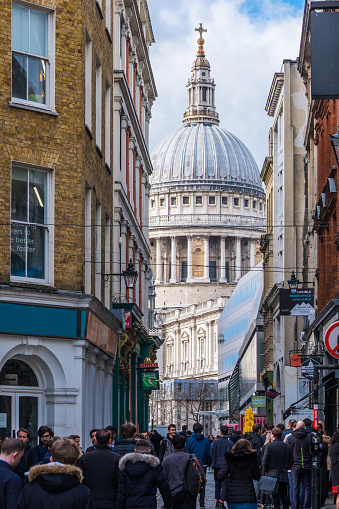 The iconic dome of St. Paul’s Cathedral between the shopfronts and restaurants in the narrow crowded streets of the City of London Financial District, UK.