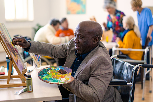 An retired Anglo-Caribbean man is painting onto a canvas, holding a palette in one hand as he reaches with his other from his seat in a wheelchair, in the background more senior people are sitting around a table in a residential care home
