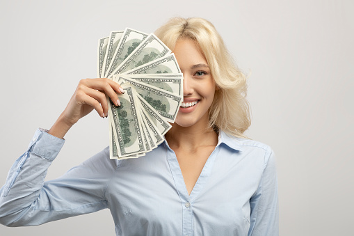 Portrait of happy business lady hiding face behind cash, holding bunch of dollars by face and smiling over light grey background. Female entrepreneur showing fan of US banknotes