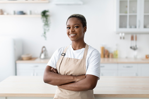 Cheerful millennial african american female in apron with crossed arms looking at camera at modern minimalist kitchen interior. Chef, food blogger, housewife at home and ad during covid-19 outbreak