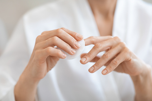 Woman hand removing nail polish with white cotton pad, unrecognizable lady in white bathrobe doing manicure for herself at home, cropped. Manicure, hands care concept