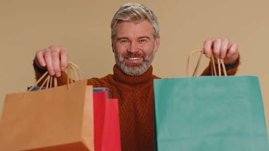 Middle-aged old man showing shopping bags, advertising discounts, smiling looking amazed with low prices, shopping on Black Friday holidays. Senior mature guy isolated alone on beige studio background