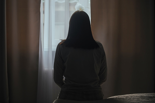 Silhouette of a young woman in pajamas against the background of a window at home on a bed with seasonal affective disorder or depression. The concept of winter depression due to lack of sunlight