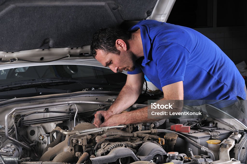 Male mechanic in blue polo shirt working on a car engine A young man with black hair, a blue polo shirt, and a tool in his right hand is bending over the right side of the open hood of an off-white car.  The engine of the car is exposed and appears slightly dusty.  The image is on a black background. Mechanic Stock Photo