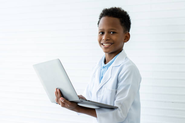 коротковолосый афроамериканский мальчик в белом лабораторном халате держит в обеих руках компьютерный ноутбук. он радостно улыбнулся, гот - chemistry chemistry class technician student стоковые фото и изображения