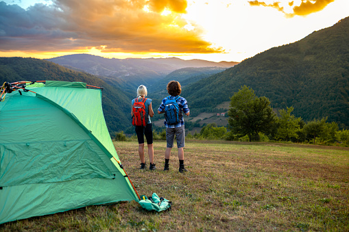 Young adult couple of hikers enjoying in nature and camping on mountain range.\nThey are watching a beautiful sunset .