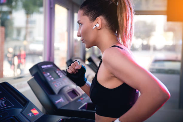 una mujer con camisetas negras y auriculares bluetooth funcionando en una banda de rodadura en un gimnasio. - run of the mill fotografías e imágenes de stock