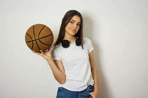 beautiful young woman holding basketball  having earphones on her neck standing over gray background