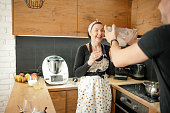 Laughing lovely family of woman in apron and rearview man holding flour bowl and fooling around against kitchen drawers