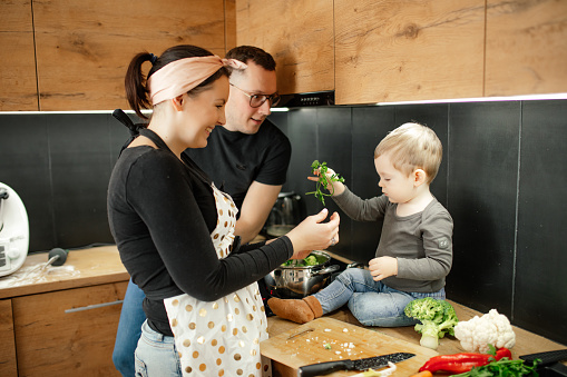 Satisfied smiling family of woman in apron, man in glasses talk and teaching little blond boy to cook in kitchen. Vegetable salad recipe, housewife chores and little helper. Parenthood responsibility