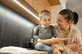 Tender laughing positive brunette woman carrying little blond boy with rolling pin in hands, cooking together in kitchen
