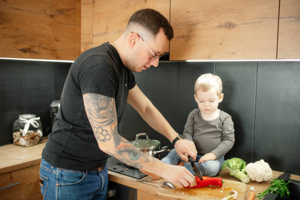 istock Father with little child cook dinner side view. Baby son sitting on tabletop help dad cut red bell pepper on board. 1468874218