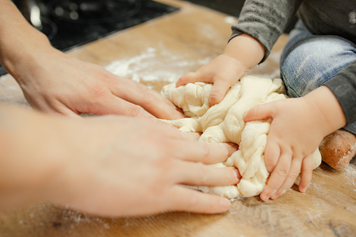 Parent with baby together knead raw elastic dough on kitchen tabletop closeup. Little hands help mom cooking dinner, flour dish. Homemade recipe, food, baking pastry. Family lifestyle.