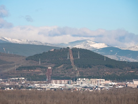 View of the city of Ponferrada where the Rosaleda tower stands out and a small mountain with a firebreak and the snowy mountains in the background with clouds around a sunny day