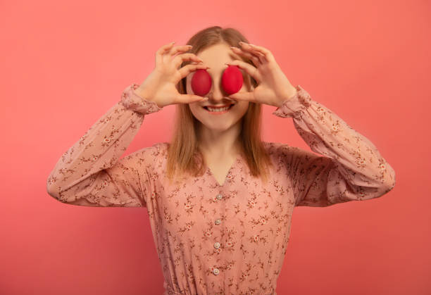 istock Happy smiling young woman holding two red painted easter eggs or two red dyed easter eggs on eyes making joyful face expression isolated on pink background.

Cheerful girl celebrating Easter holiday. 1468883840