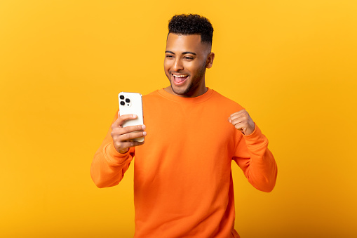 Happy satisfied man holding smartphone and smiling making yes gesture, celebrating online lottery or giveaway victory. Indoor studio shot isolated on orange background