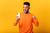 Happy satisfied man holding smartphone and smiling making yes gesture, celebrating online lottery or giveaway victory. Indoor studio shot isolated on orange